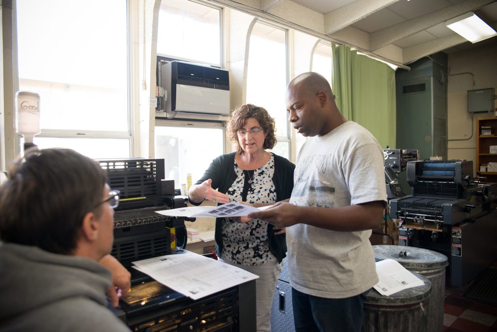 Kathy Harris, a white woman in a black and white blouse, looks over classwork with student Maurice Hollins, a black man in a grey t-shirt.