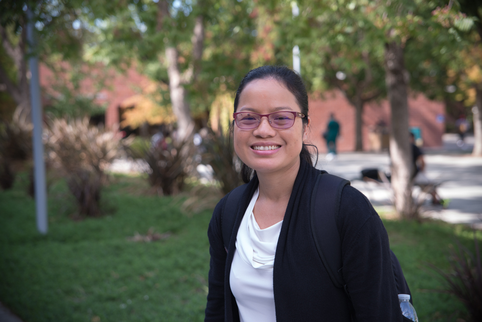 Somying Hughes, a Thai woman in glasses and a white shirt with a black jacket and backpack, stands outside at her adult school campus.