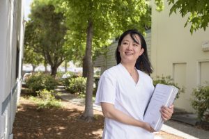 Yan Chen stands outside in white scrubs holding school notes and books.