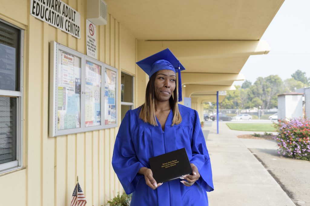 Angela Byrd stands outside a building wearing her graduation cap and gown and holding her high school equivalency diploma.