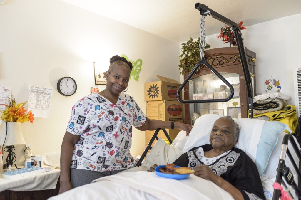 Vanessa Fines in scrubs stands at the bedside of an elderly male patient in his home.