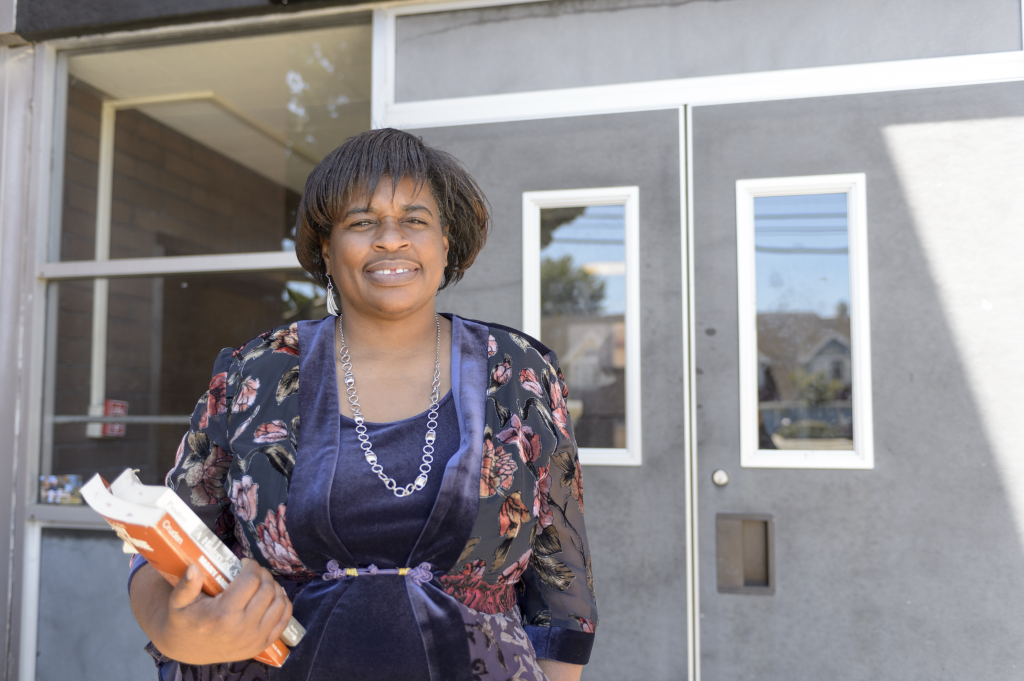 A woman who did not finish high school stands in front of a pair of double doors. She is holding a stack of books and smiling at the camera. 