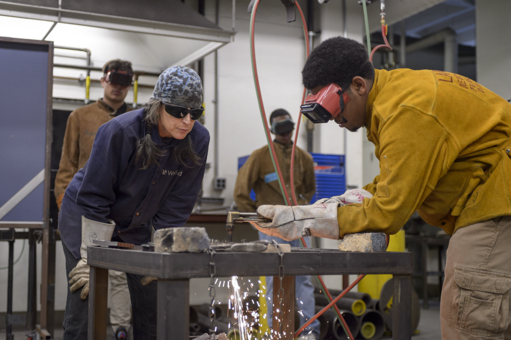 An older woman and a young man are welding. They are both wearing protective eye wear. There are a few people watching them from behind. 