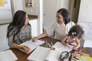 A mother and her two children sit at a table in their home, smiling over textbooks and pencils.