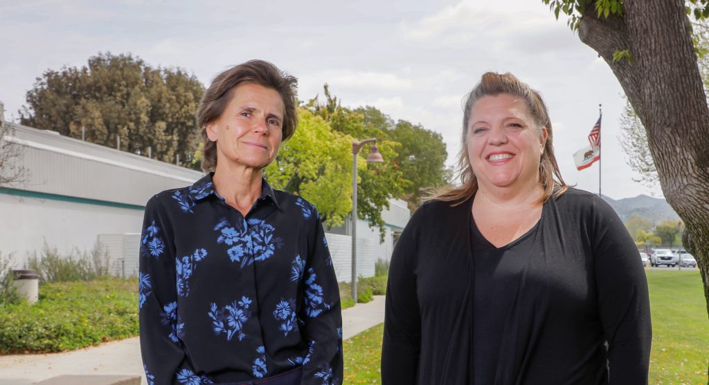 Ute Maschke (left) and Sally Cox (right) stand outside an adult education college and smile for the camera.