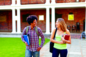 Two young students, one male and one female, on campus holding books smiling at each other.