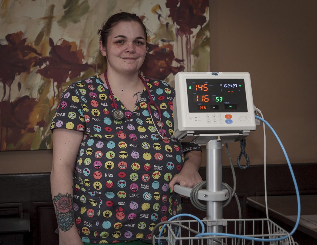 Raven Ortega, wearing colorful scrubs, standing beside medical monitoring equipment.