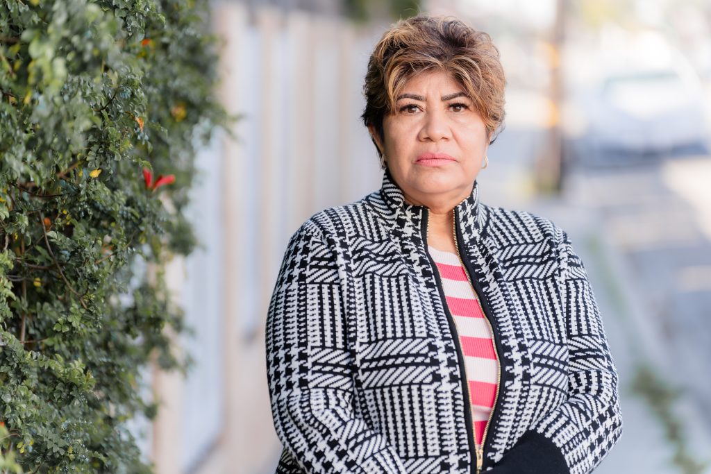 Maria Rodriguez, a woman with short hair and a black and white houndstooth jacket, stands outside and looks towards the camera.