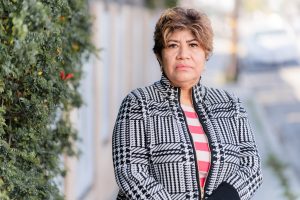 Maria Rodriguez, a woman with short hair and a black and white houndstooth jacket, stands outside and looks towards the camera.