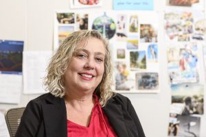 Linette Escobar, a woman with short, wavy blond hair, a pink blouse, and a black blazer smiles for the camera from her office