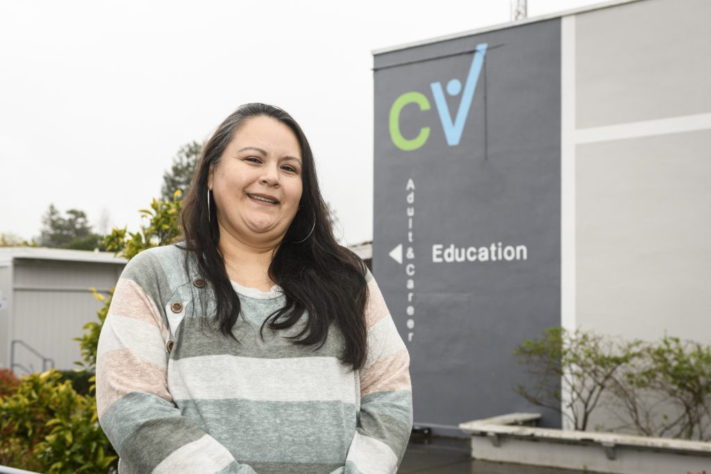 Iema Becerra, a woman with long straight brown hair and a striped long sleeves shirt, stands in front of the CVACE building.