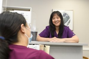 Julie Siroy, an Asian woman wearing purple scrubs, leans on a nurse's desk while smiling at a coworking