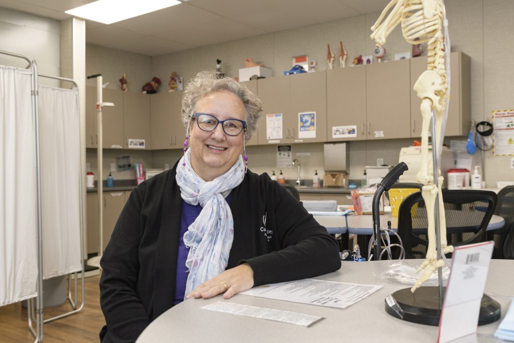 Elisa Dasalla, a woman with short grey hair, a scarf, and a black shirt, sits in her classroom and smiles for the camera