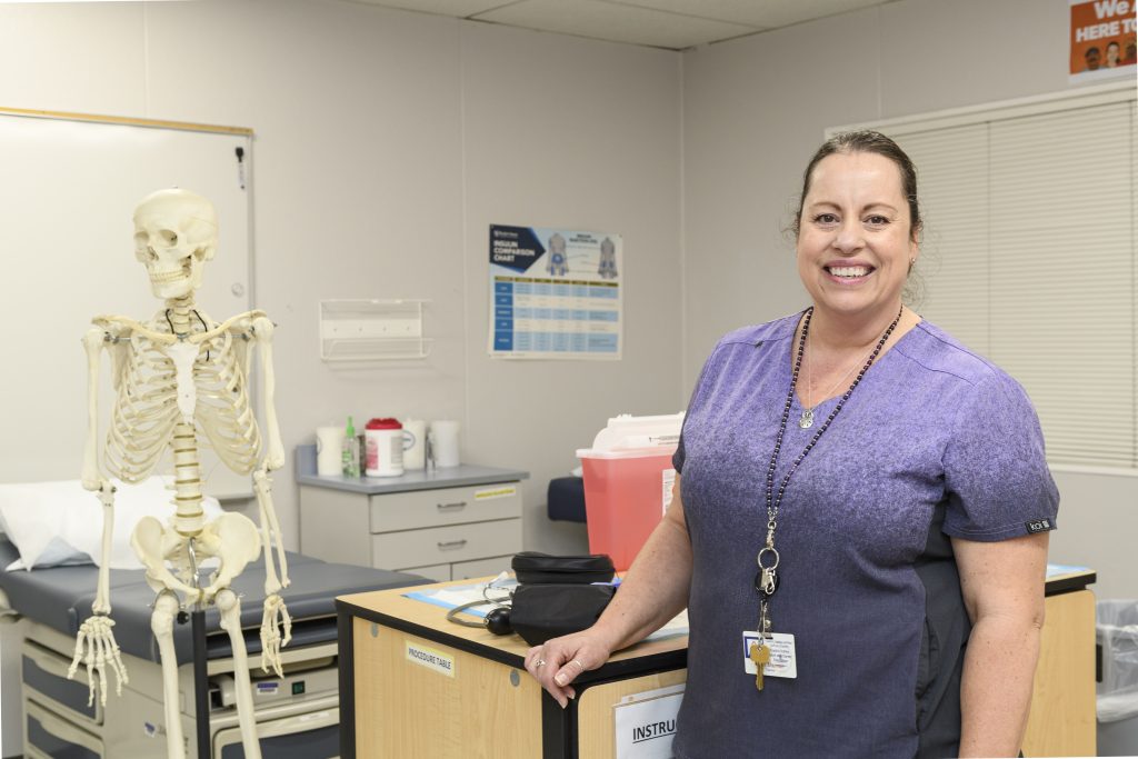 Shelli Sherman, a woman with her brown hair pulled back and wearing purple scrubs, stands in her classroom and smiles for the camera.