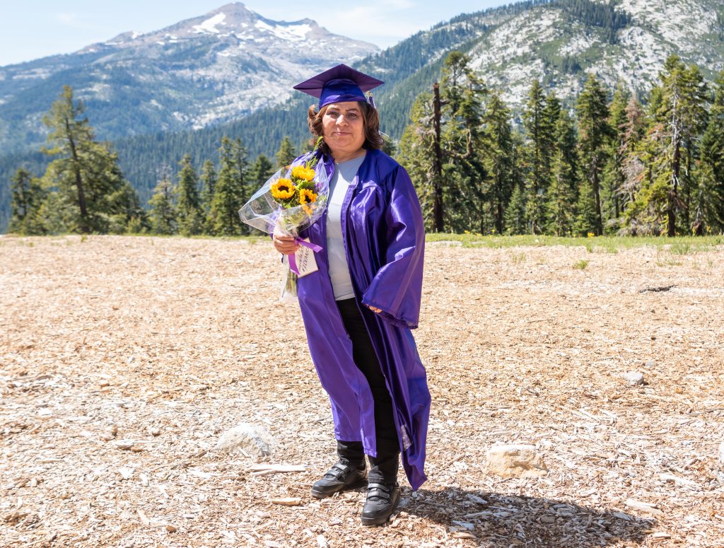 Hermalinda Aguirre, wearing a purple graudation cap and gown and holding a bouquet of sunflowers, stands in front of a landscape of pine trees and mountains