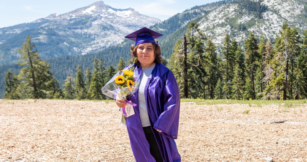 Hermalinda Aguirre, wearing a purple graudation cap and gown and holding a bouquet of sunflowers, stands in front of a landscape of pine trees and mountains