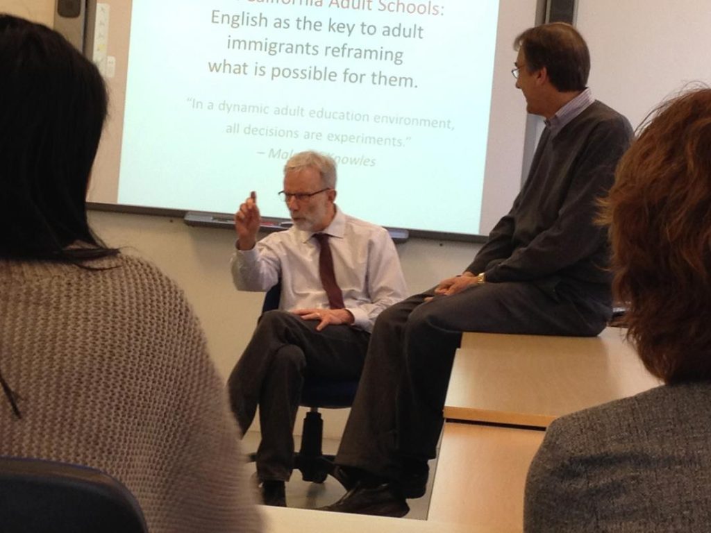 Bob Harper, Former executive director of Adult Education, South Bay Consortium for Adult Education, sits in a chair in front of a powerpoint and speaks to a classroom.