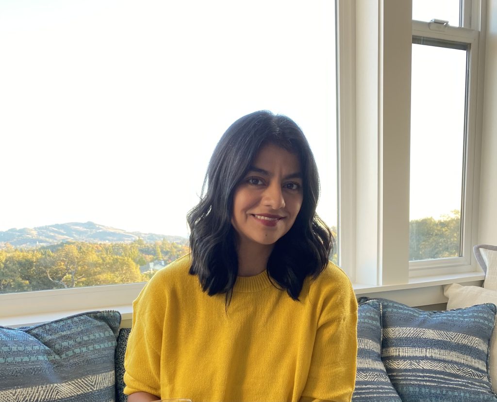 Maria Vela, a woman with shoulder length dark hair in a yellow blouse, sits on her couch and smiles for the camera