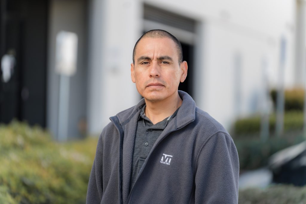 Miguel Ramirez, a man with close cropped hair, a grey polo shirt, and a grey jacket, stands outside and looks towards the camera.