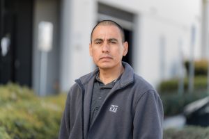 Miguel Ramirez, a man with close cropped hair, a grey polo shirt, and a grey jacket, stands outside and looks towards the camera.