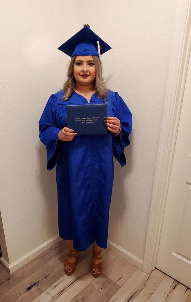 Candy Torres, a woman with brown hair and blonde highlights, smiles for the camera in her home while wearing her blue graduation cap and gown and holding her diploma.