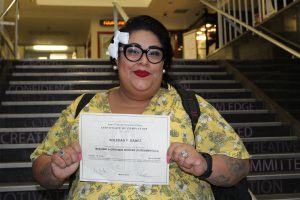 A woman in a yellow blouse with a flower in her hair and glasses holding up a certificate from an adult school