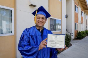 Portrait of Andres Arias, a recent graduate of Escondido Adult School wearing his cap and gown and holding his diploma in front of his southest San Diego area apartment building.