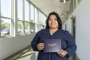 Romelia Perez, a woman with mid-length dark brown hair and a navy blouse, holds her diploma and smiles for the camera.