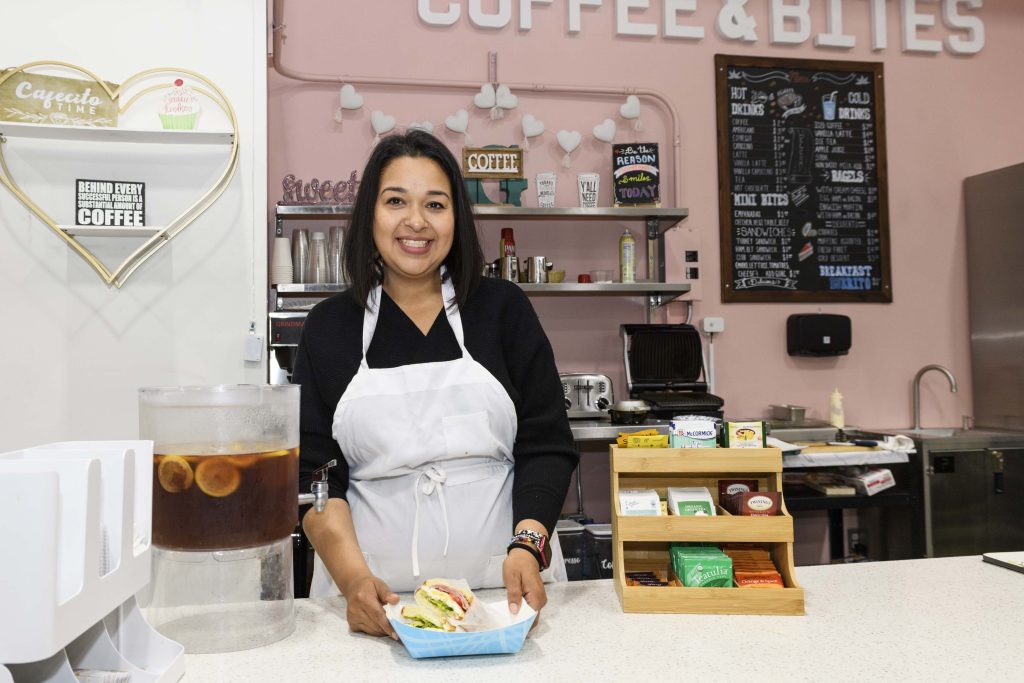 Gabby Ramirez smiles for the camera in a cafe, wearing a black shirt and a white apron and holding a freshly made sandwich