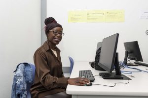 Fatima Adcock, a black woman with a brown button up shirt, glasses, and her hair up in a bun, sits at a computer in a classroom and smiles for the camera.