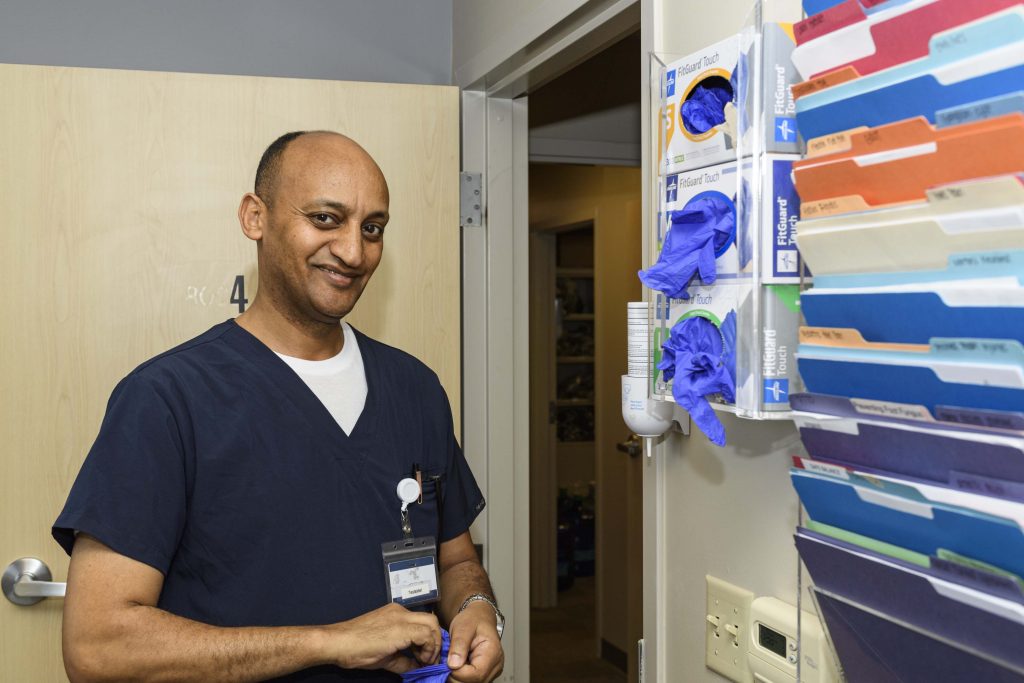 Tes Yohannes in scrubs starts to put on a latex glove and smiles for the camera