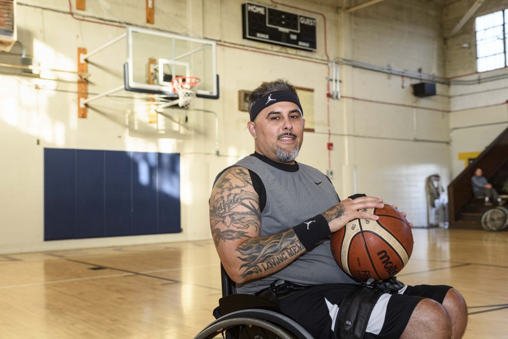 Armando Rodriguez, a man with tattoos and a gray workout shirt in a wheelchair, holds a basketball in a gym and looks towards the camera