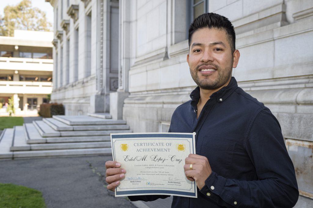 Edel Lopez stands outside holding his GED and smiles for the camera