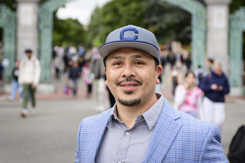 Juan Flores, in a light blue ball cap and blazer with a light grey button up, smiles for the camera