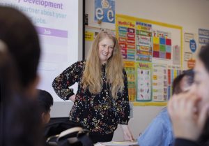 Polina Trask, a woman with long blonde hair and a black blouse with a multi-colored pattern, teaches in her adult education classroom.