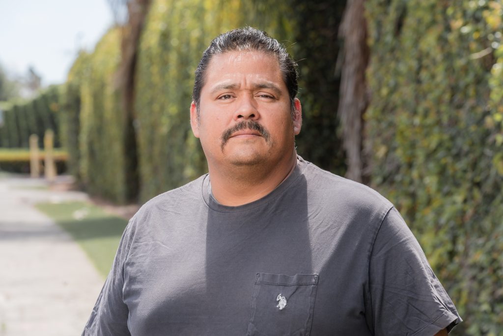 Orlando Ayala, a Latino man with a mustache and a gray shirt, stands outside in front of tall hedges and looks towards the camera