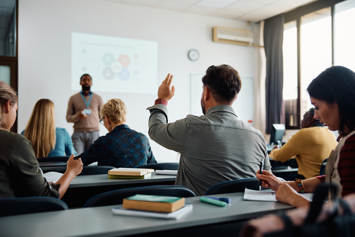 Rear view of man raising arm to ask a question during a presentation in lecture hall.