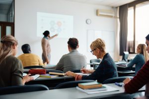 Several adults from the back sitting in a classroom watching a teacher lecture