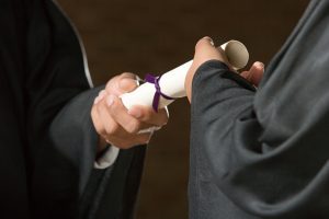 A close-up of two people in academic gowns, one handing a rolled diploma to the other