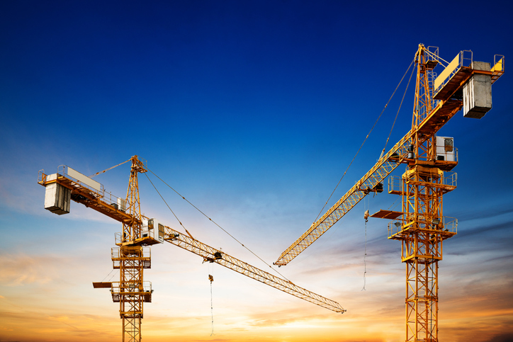 Industrial construction cranes and building silhouettes over sun at sunrise.