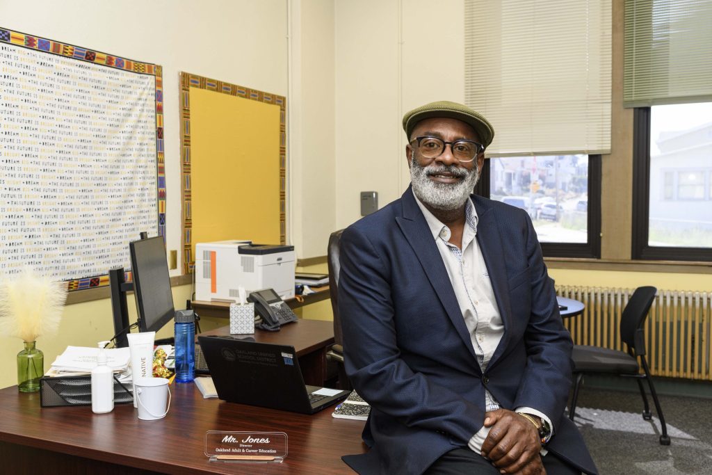 Kim Jones, an older black man with a gray beard, blazer, and flat cap, sits on the desk in his office and smiles for the camera