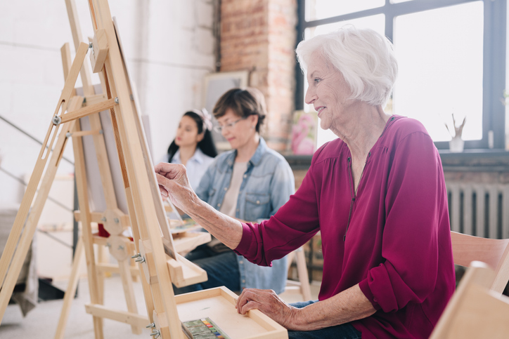 Side view portrait of art students sitting in row and painting at easels in art studio, focus on smiling elderly woman