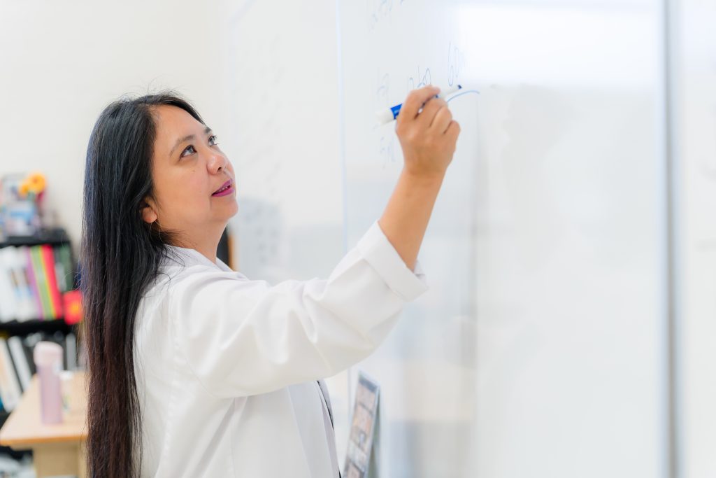 Leticia Wang in a lab coat writing on a whiteboard in a classroom