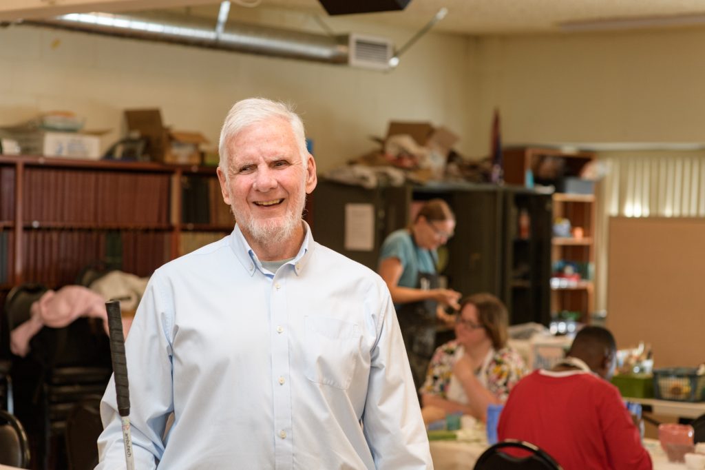 Mike Cole, a man with white hair and beard in a white button up shirt, stands in a classroom at the East Bay Center for the Blind and smiles for the camera