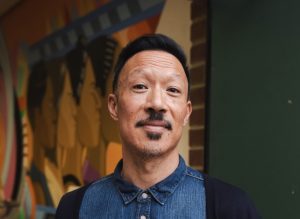 Roger Chung, a man with a mustache and small goatee wearing a demin shirt, stands and smiles for the camera