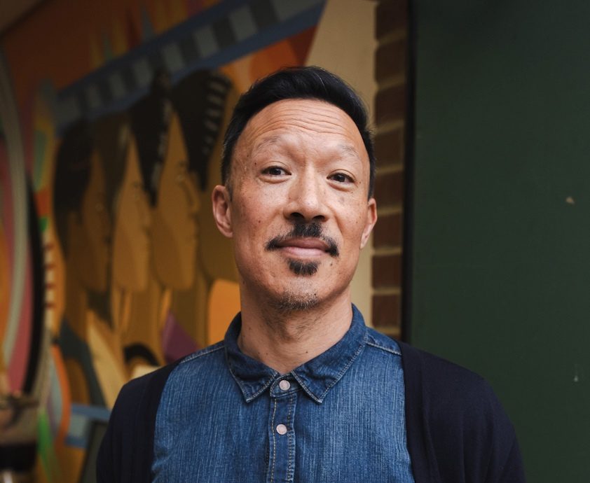 Roger Chung, a man with a mustache and small goatee wearing a demin shirt, stands and smiles for the camera