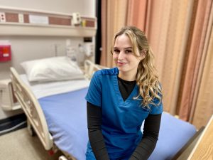 Bek Grey in blue scrubs sitting on the edge of a hospital bed