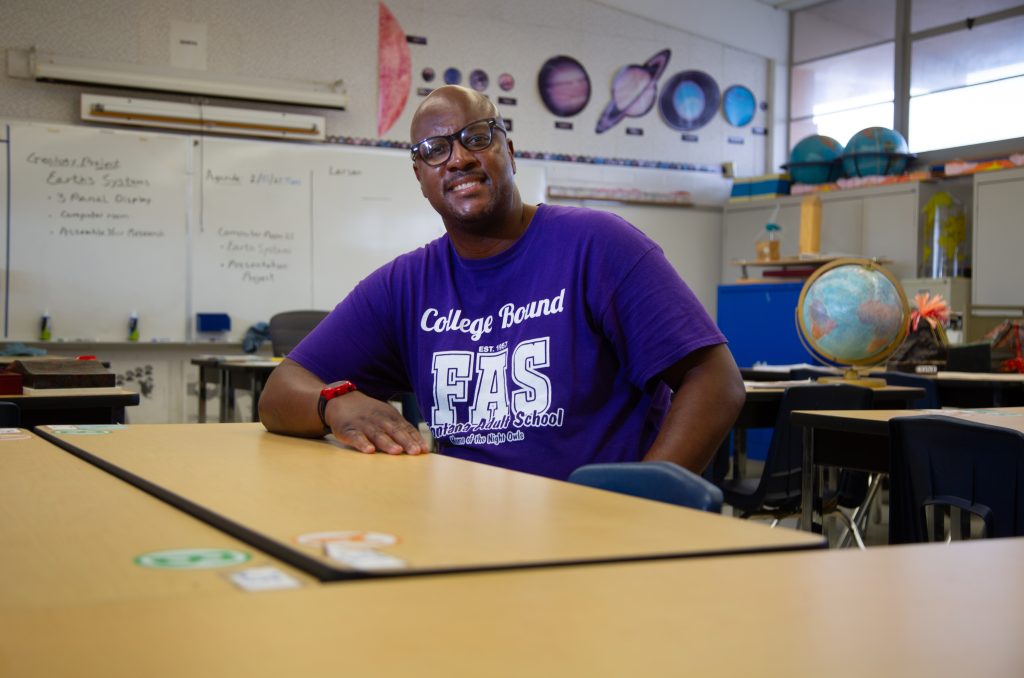 Man smiling and sitting at a school desk in a classroom.
