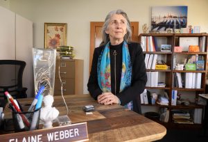 Elaine Webber in black zip up jacket and blue scarf stands at her office desk