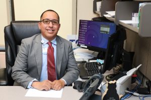 Oscar Morales in a grey suit with a red tie sitting in an office at a computer desk